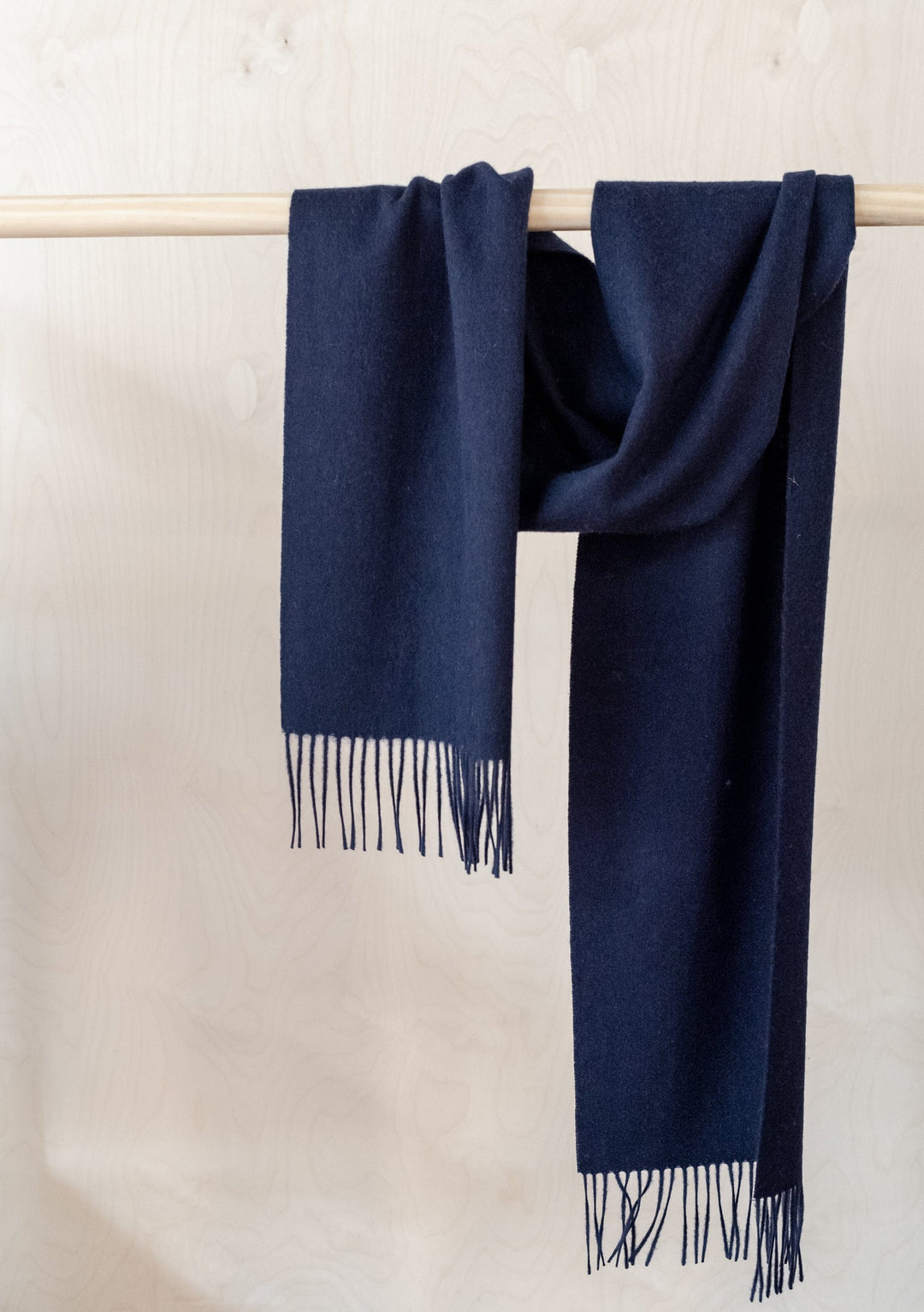 Luxurious navy TBCo lambswool scarf with fringed ends draped elegantly over wooden rail displaying soft texture and rich colour