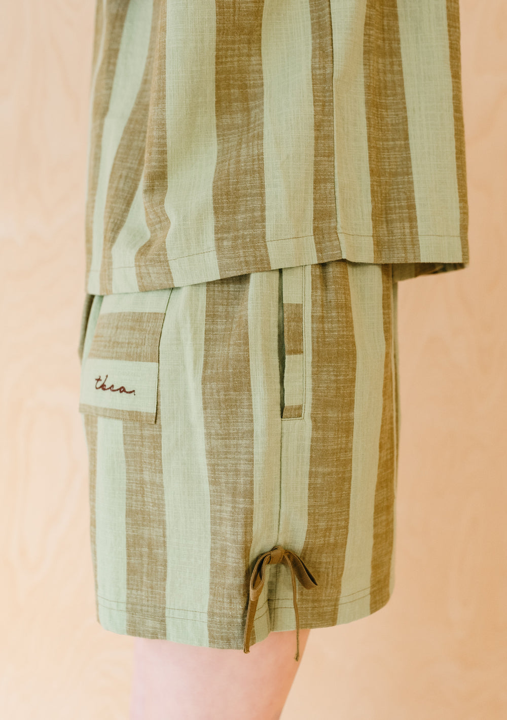 Side view of TBCo Cotton Tie Shorts in Green & Brown Stripe featuring drawstring hem and patch pocket detail on light background