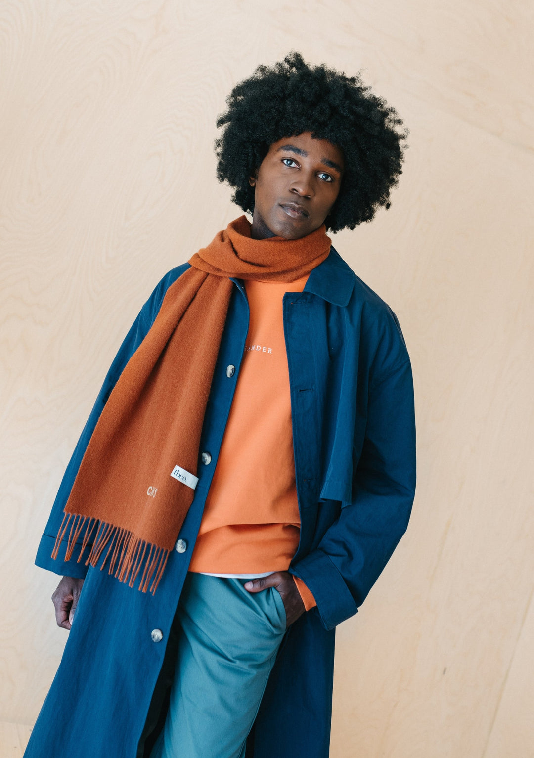 A person wearing TBCo's rust melange lambswool scarf styled with a navy coat and orange jumper against a beige background