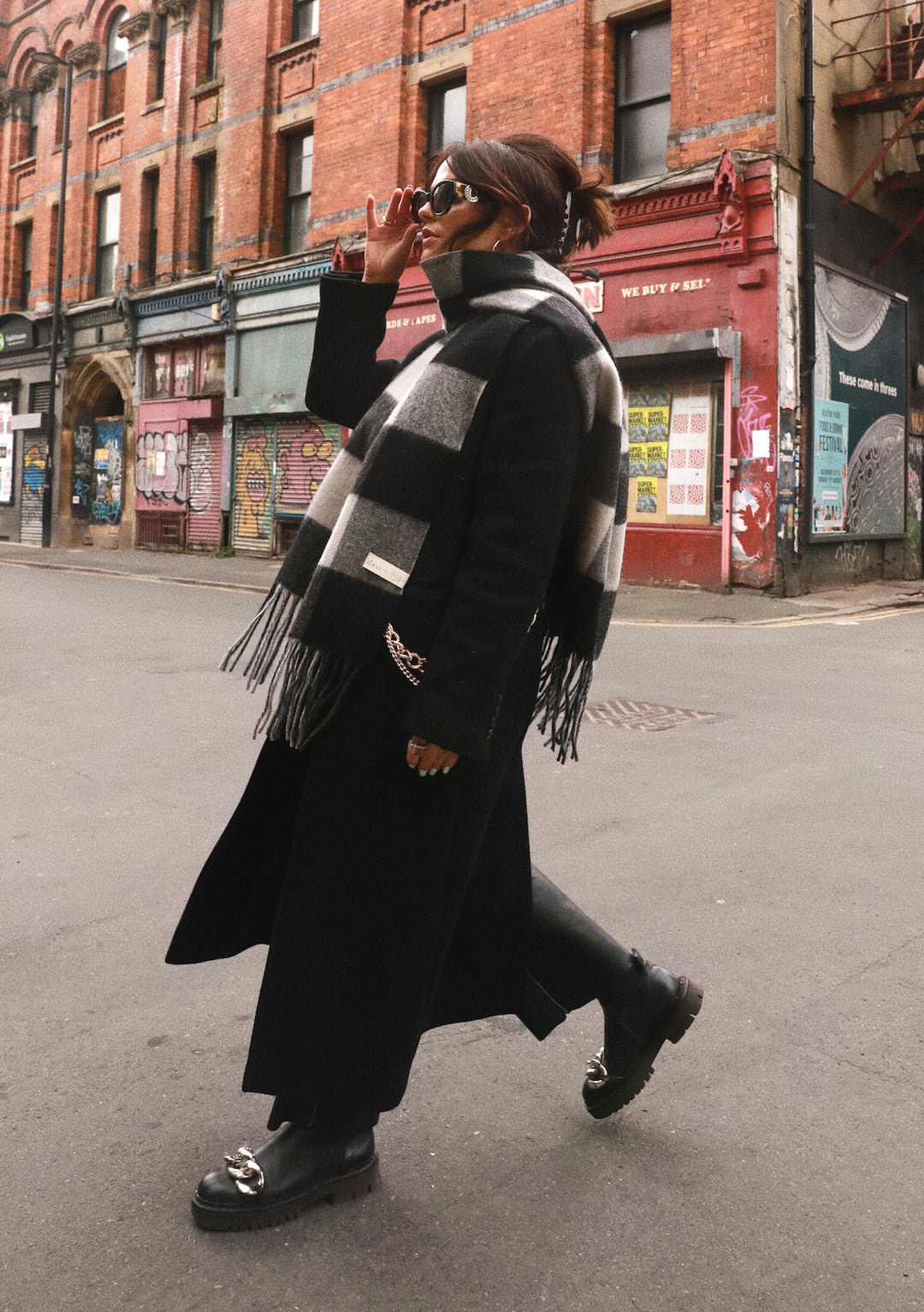 A person wearing TBCo x Amy Bell Spencer 2.0 scarf in monochrome check pattern with black winter coat on urban street
