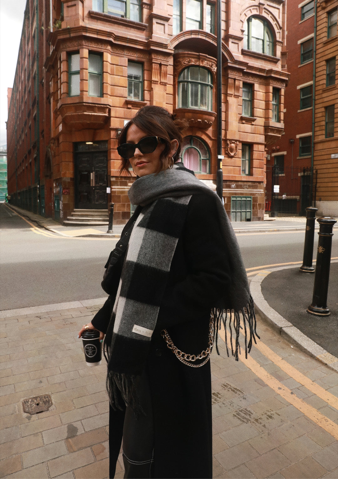 A person wearing TBCo x Amy Bell Spencer 2.0 scarf in monochrome check pattern against urban brick architecture in Edinburgh