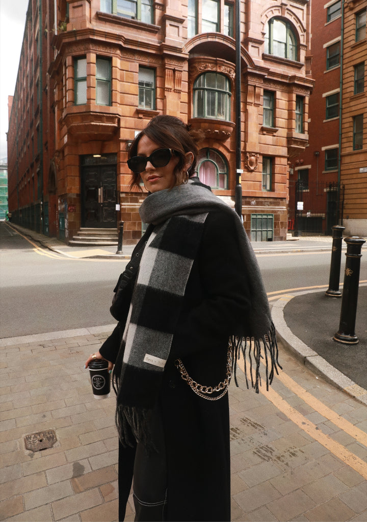 A person wearing TBCo x Amy Bell Spencer 2.0 scarf in monochrome check pattern against urban brick architecture in Edinburgh