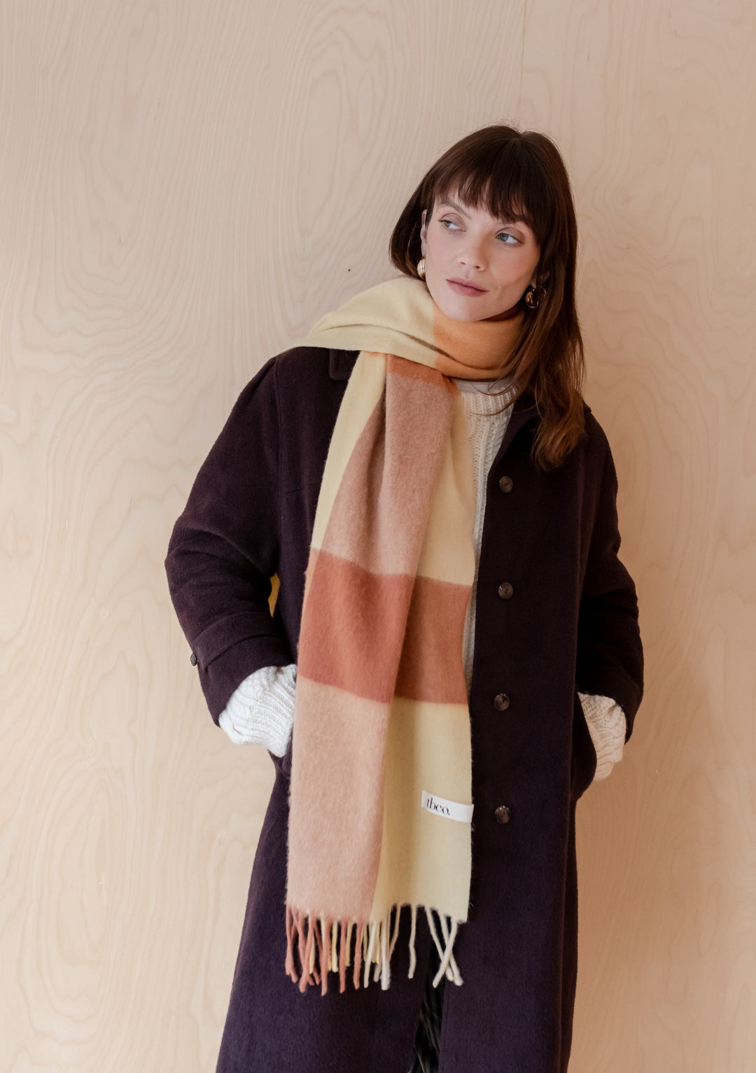 TBCo Lambswool Oversized Scarf in buttermilk yellow and rust brown check pattern, styled with dark winter coat against wooden wall