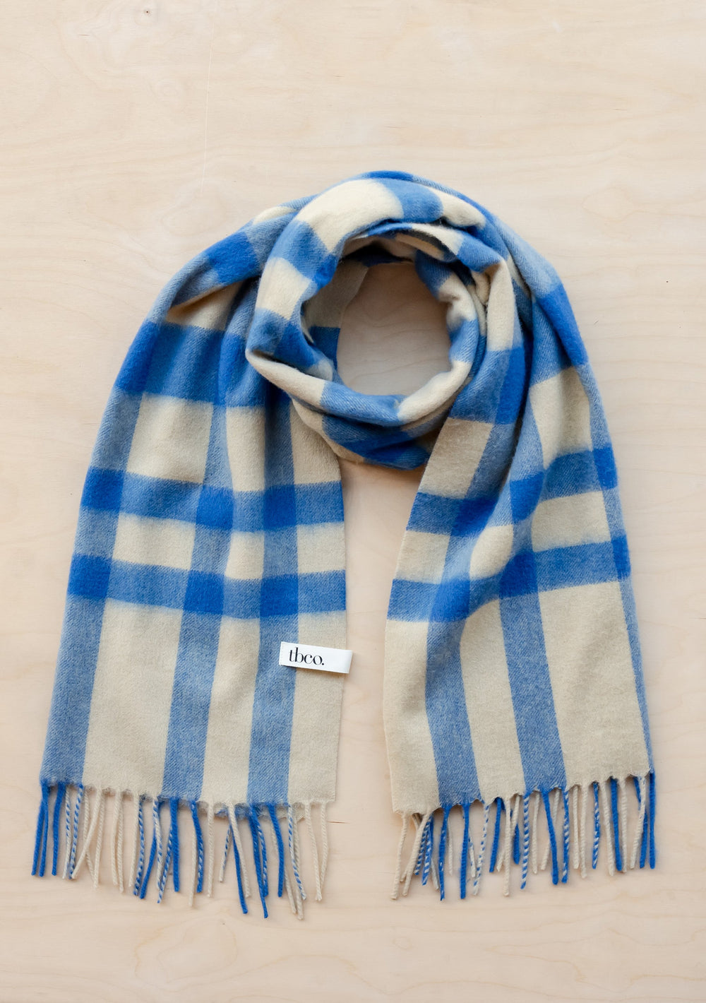 TBCo Lambswool Oversized Scarf in blue and cream check pattern with fringed edges, displayed flat on beige surface