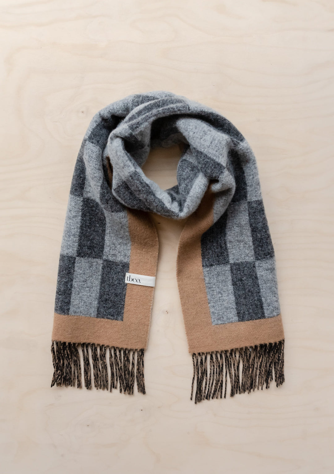 TBCo lambswool oversized scarf in grey and camel checkerboard pattern with fringed edges, laid flat on beige surface