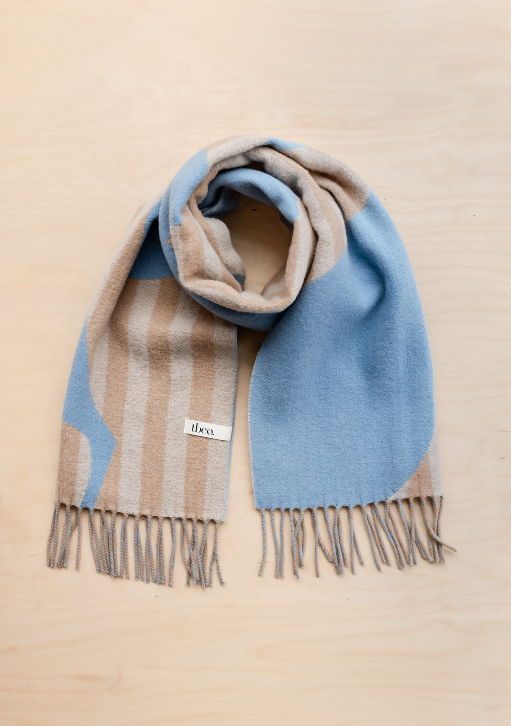 TBCo Lambswool Oversized Scarf in Flora Stripe Jacquard featuring blue and camel stripes with fringe detail on wooden surface