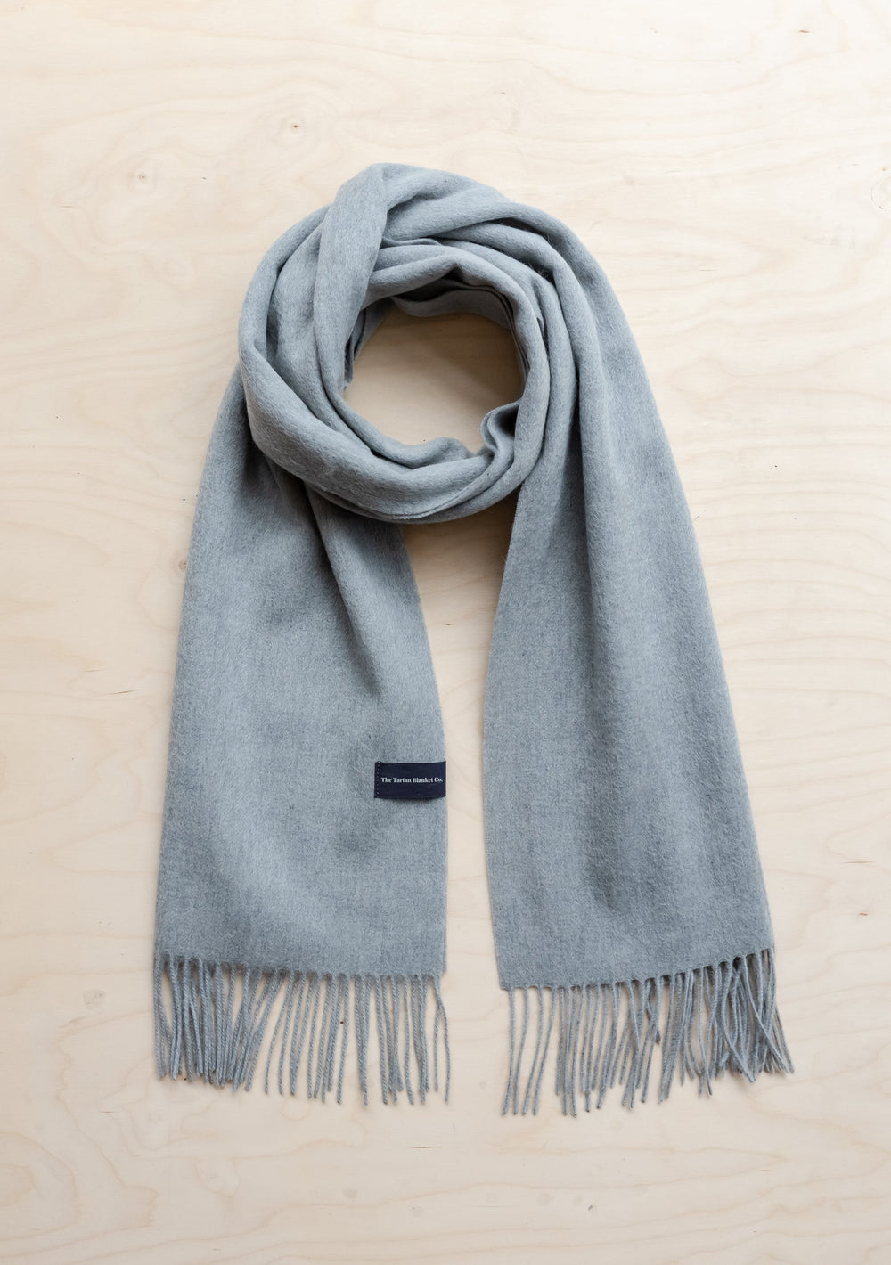 TBCo Men's Cashmere Scarf in Light Grey Melange with fringed ends, elegantly draped on light wooden surface