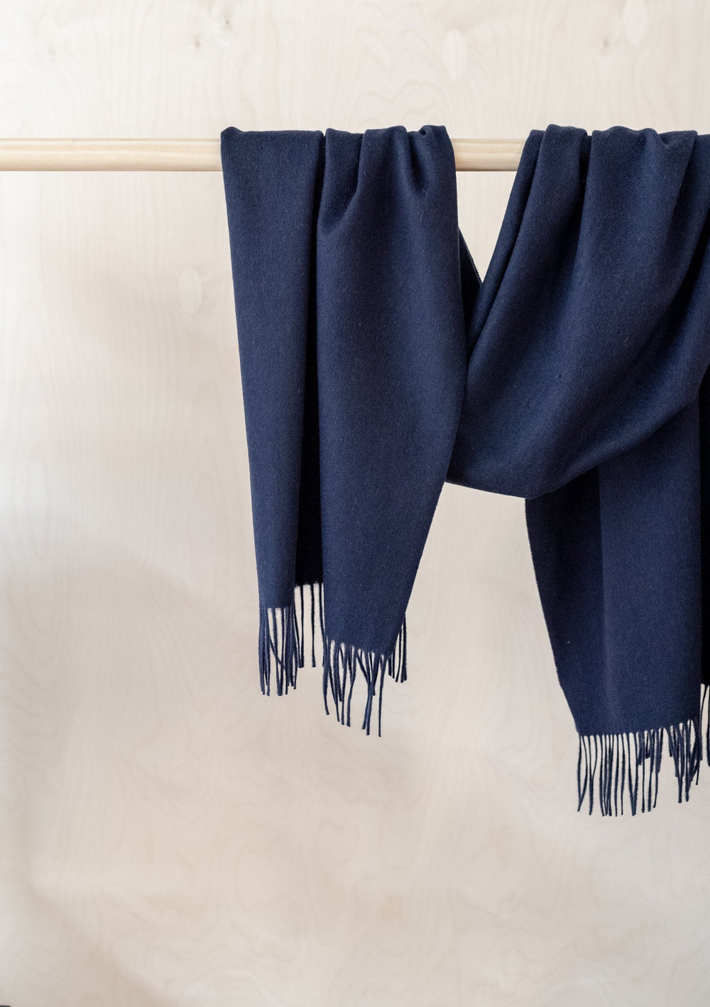 TBCo navy lambswool oversized scarf with fringe detail draped elegantly over wooden rail against white wall