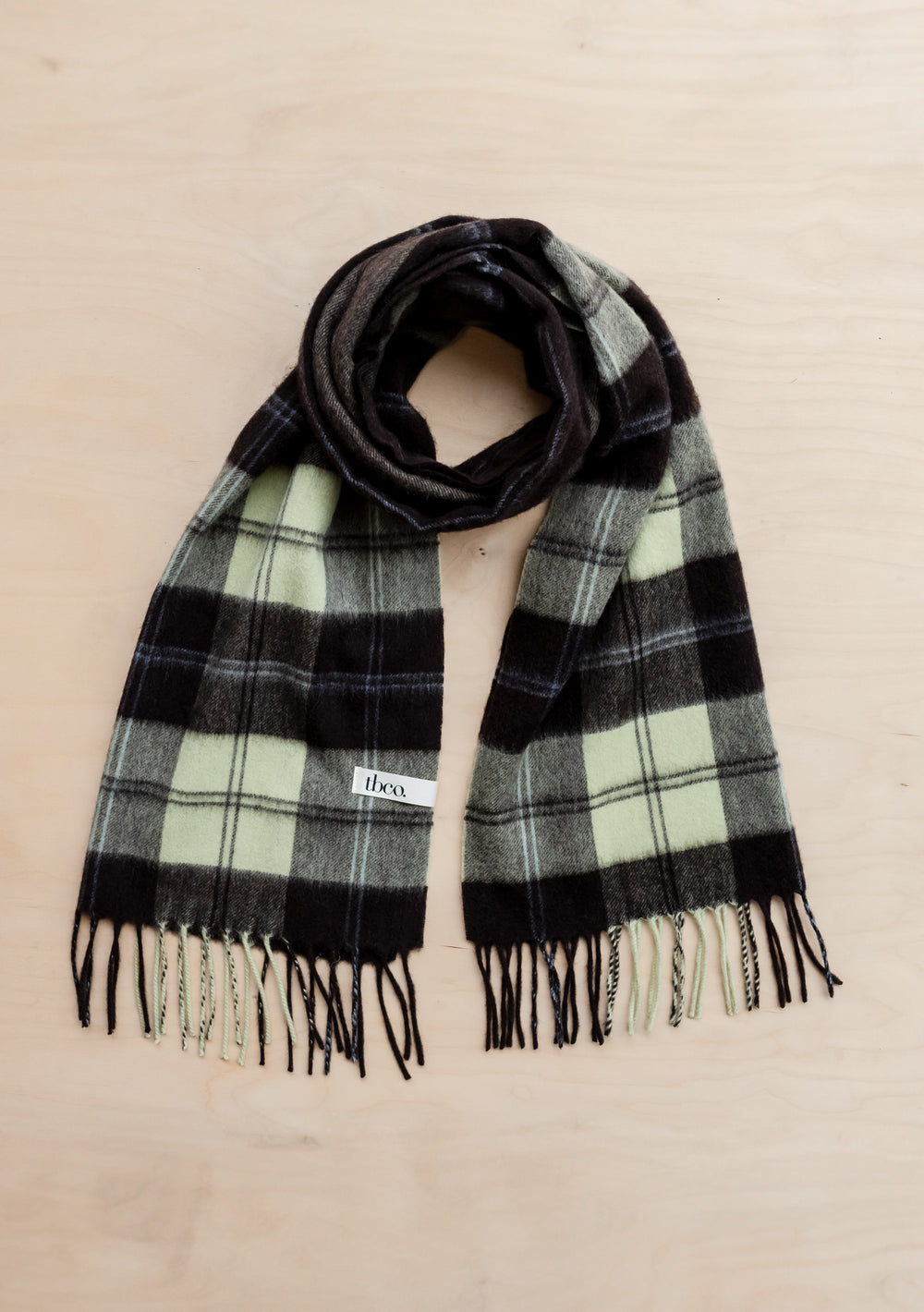 TBCo Lambswool Oversized Scarf in mint and black varsity check pattern with fringed ends, laid flat on wooden surface