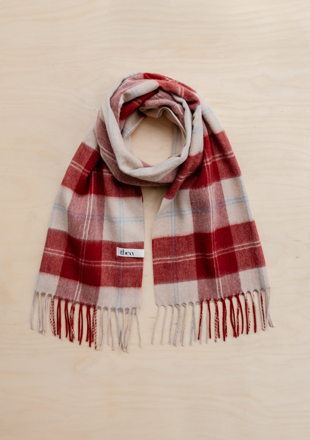 TBCo Lambswool Oversized Scarf in red and cream varsity check pattern with blue accents and fringed ends, laid flat on beige surface