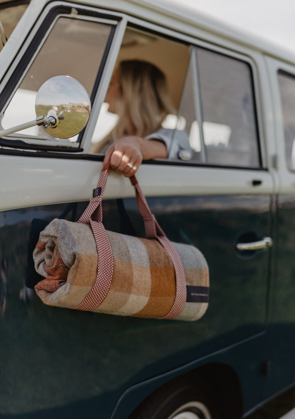 TBCo Recycled Picnic Carrier shown with vintage van, featuring plaid blanket rolled in carrier with red straps for outdoor adventures