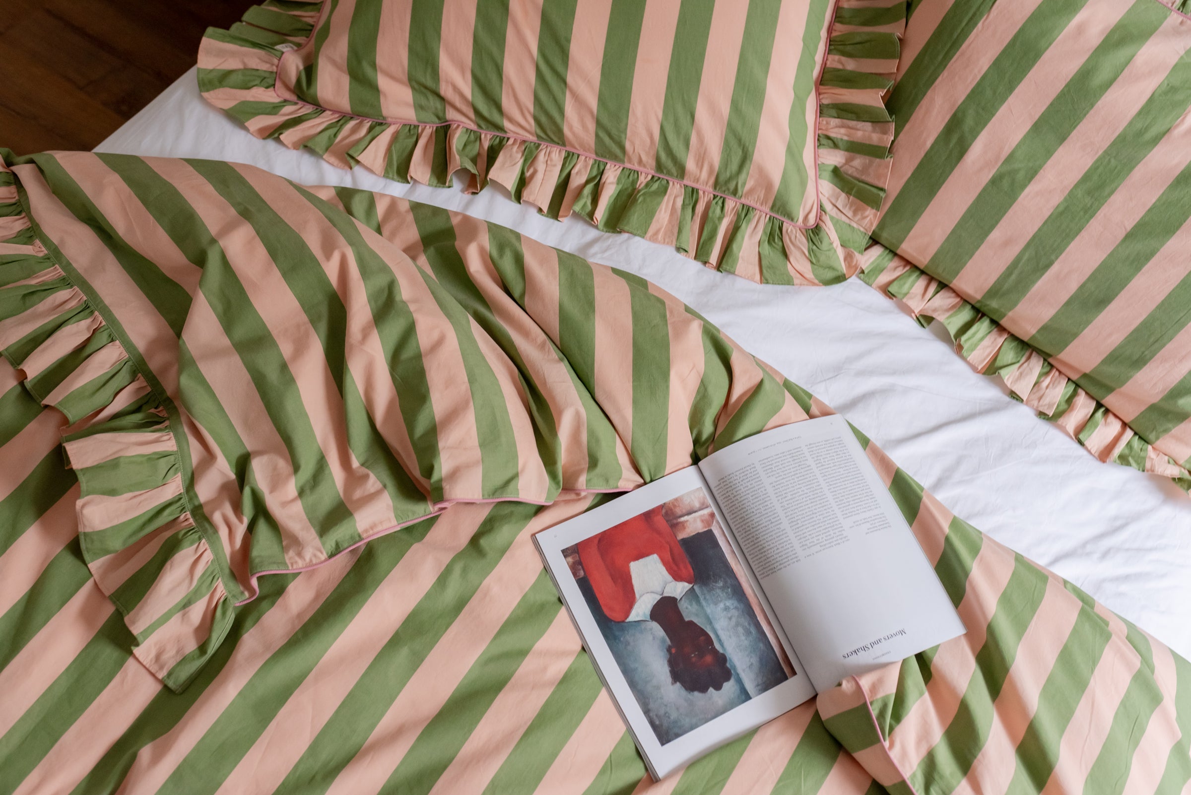 TBCo green stripe duvet cover bedding with ruffled pillowcases styled on a bed with white sheets, featuring an open magazine for a relaxed, design-forward bedroom look.