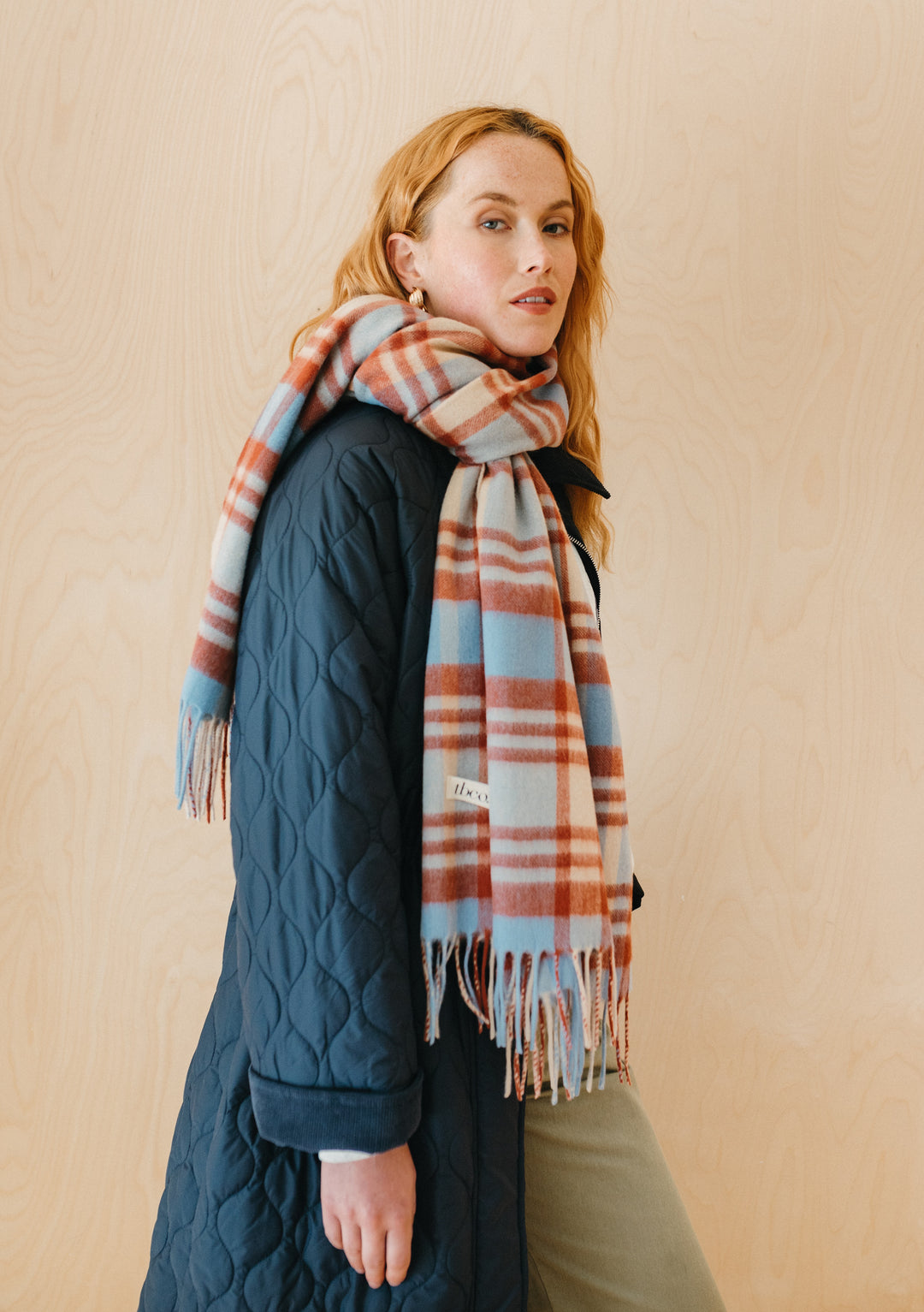 Model wearing TBCo Lambswool Blanket Scarf in Blue Tartan Check, styled with navy quilted coat against beige background