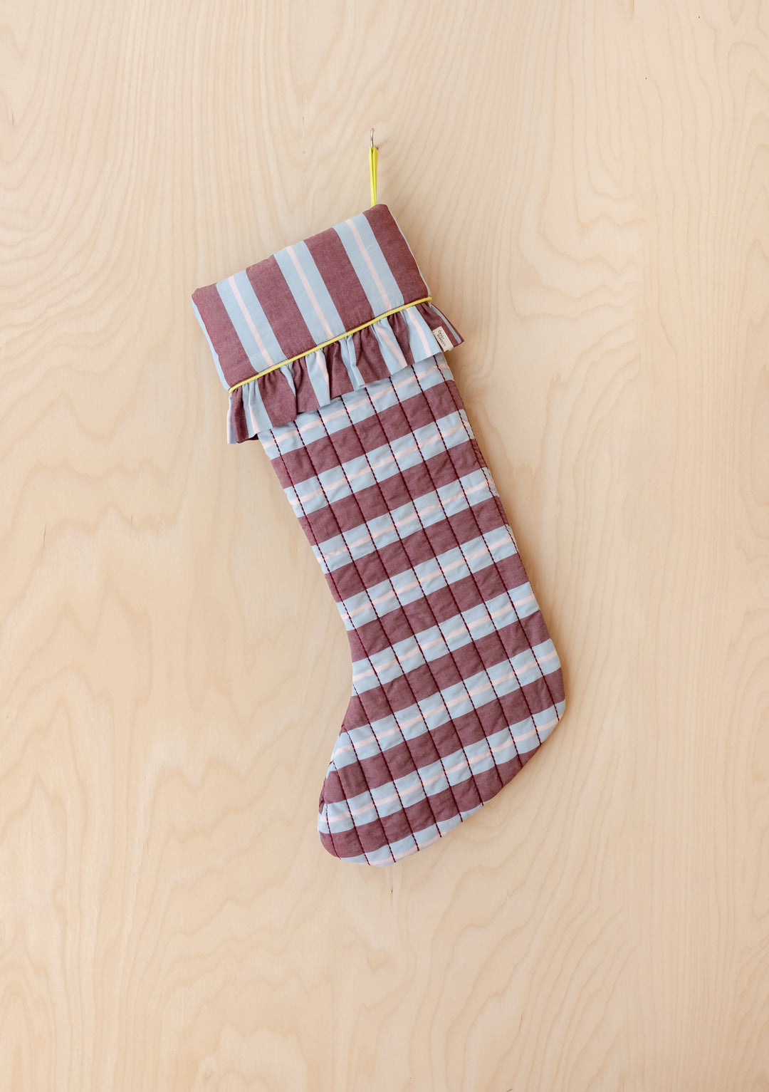 TBCo x Glassette Quilted Cotton Christmas Stocking in Berry Stripe features bold burgundy and blue stripes, ruffled cuff, and lime piping, hanging on a wood-grain wall.