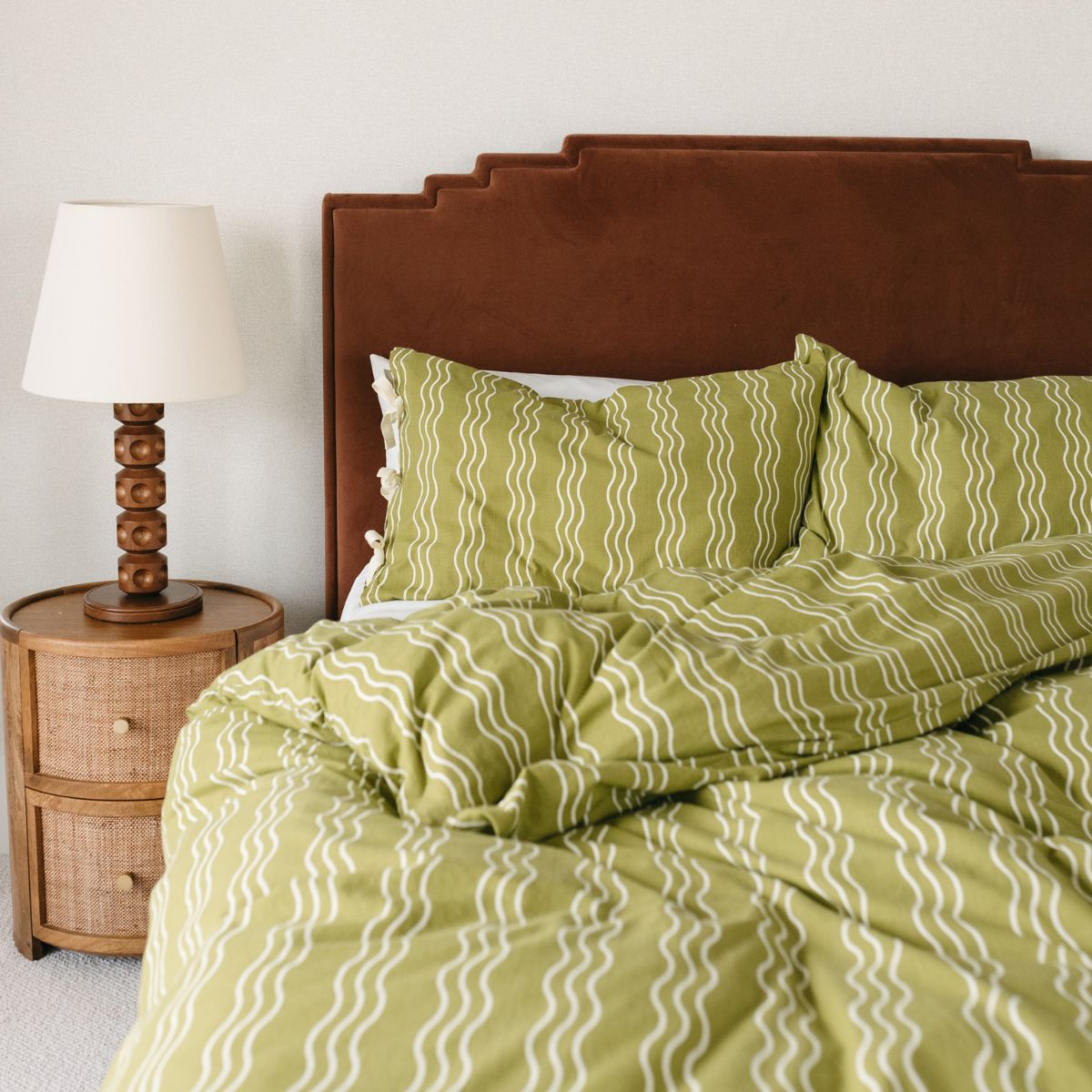 TBCo olive green cotton bedding with white wavy stripes, styled on a bed with a rust velvet headboard and wooden bedside table. A cosy and modern bedroom aesthetic.