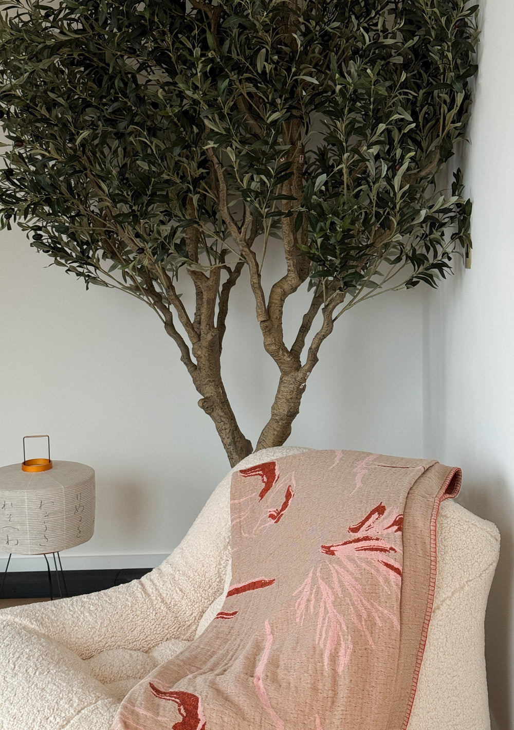 TBCo x Abisola Omole Reversible Wilderness Cotton Throw with tan base and red floral pattern, draped over chair under indoor tree