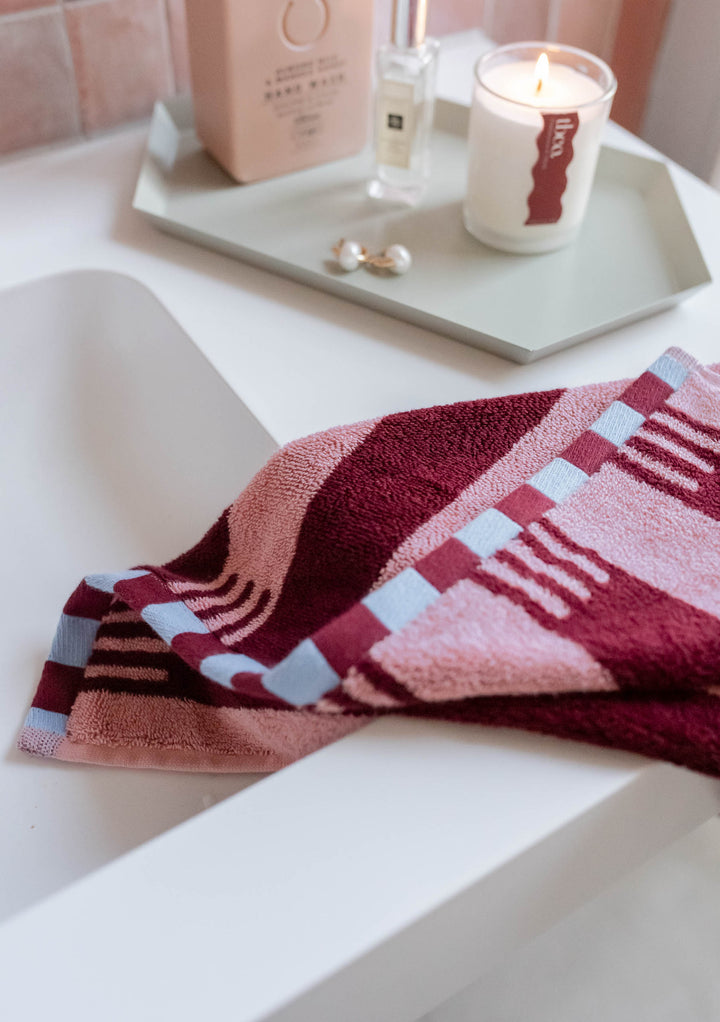 TBCo Terry Cotton Towels in Burgundy Stripe draped over bathtub edge with pink, burgundy, and blue checkerboard accents