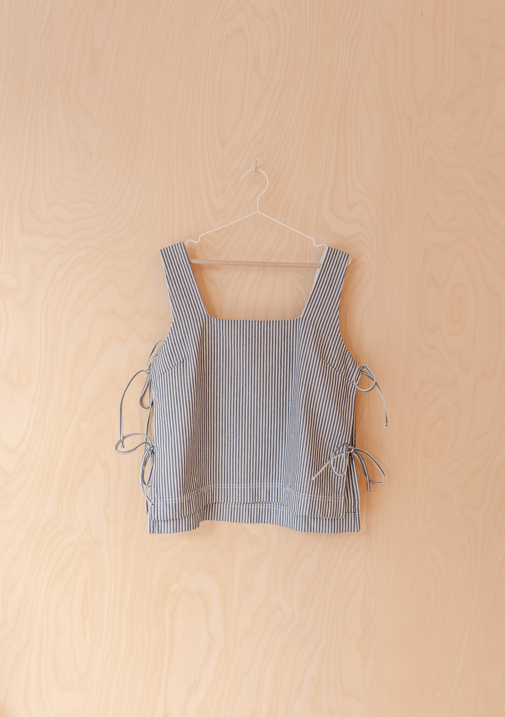 Denim Side Tie Top in Herringbone Stripe by TBCo with square neckline and side ties hanging against pale wooden wall