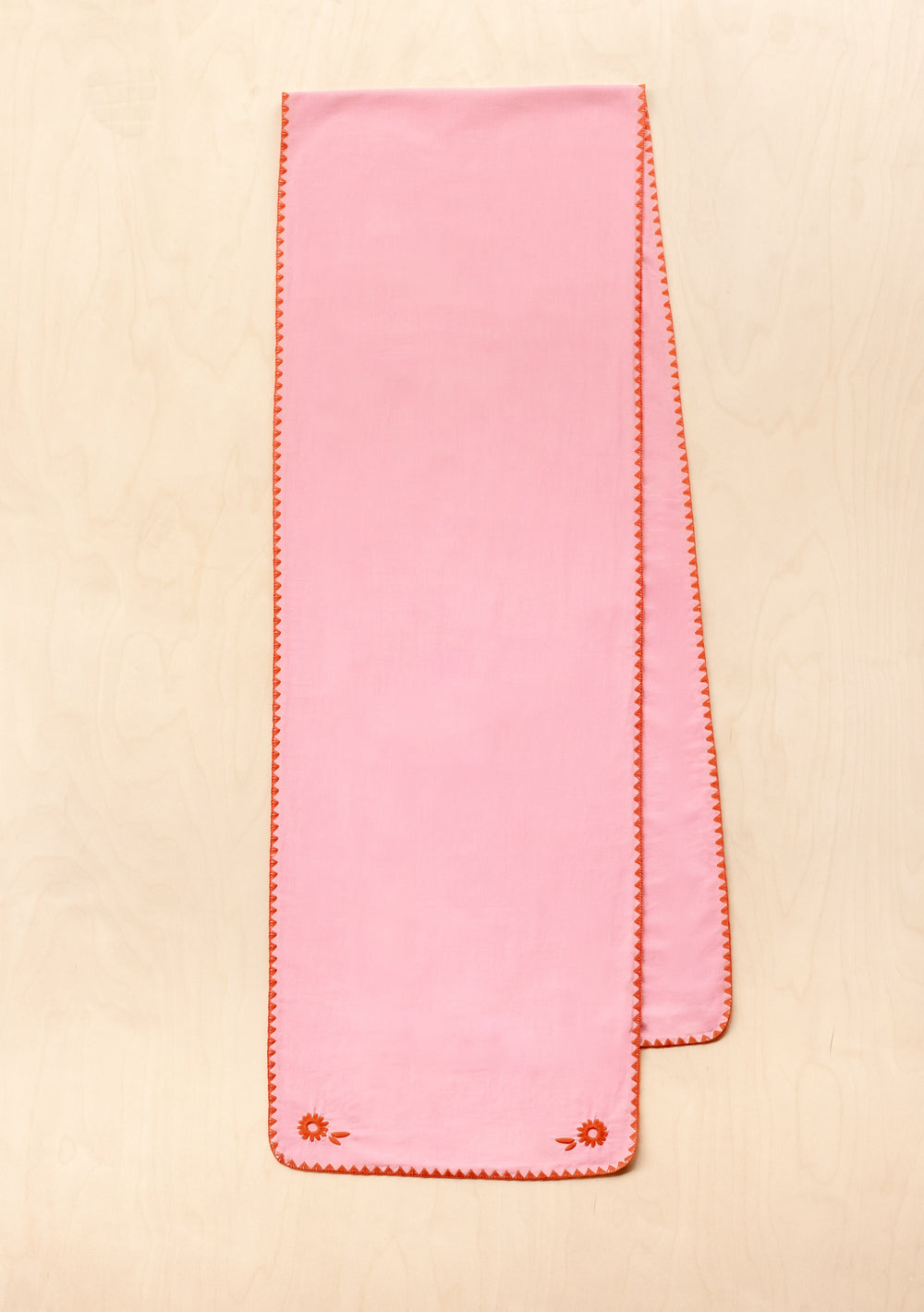 Pink cotton and linen TBCo table runner with orange floral embroidered edges displayed on wooden surface