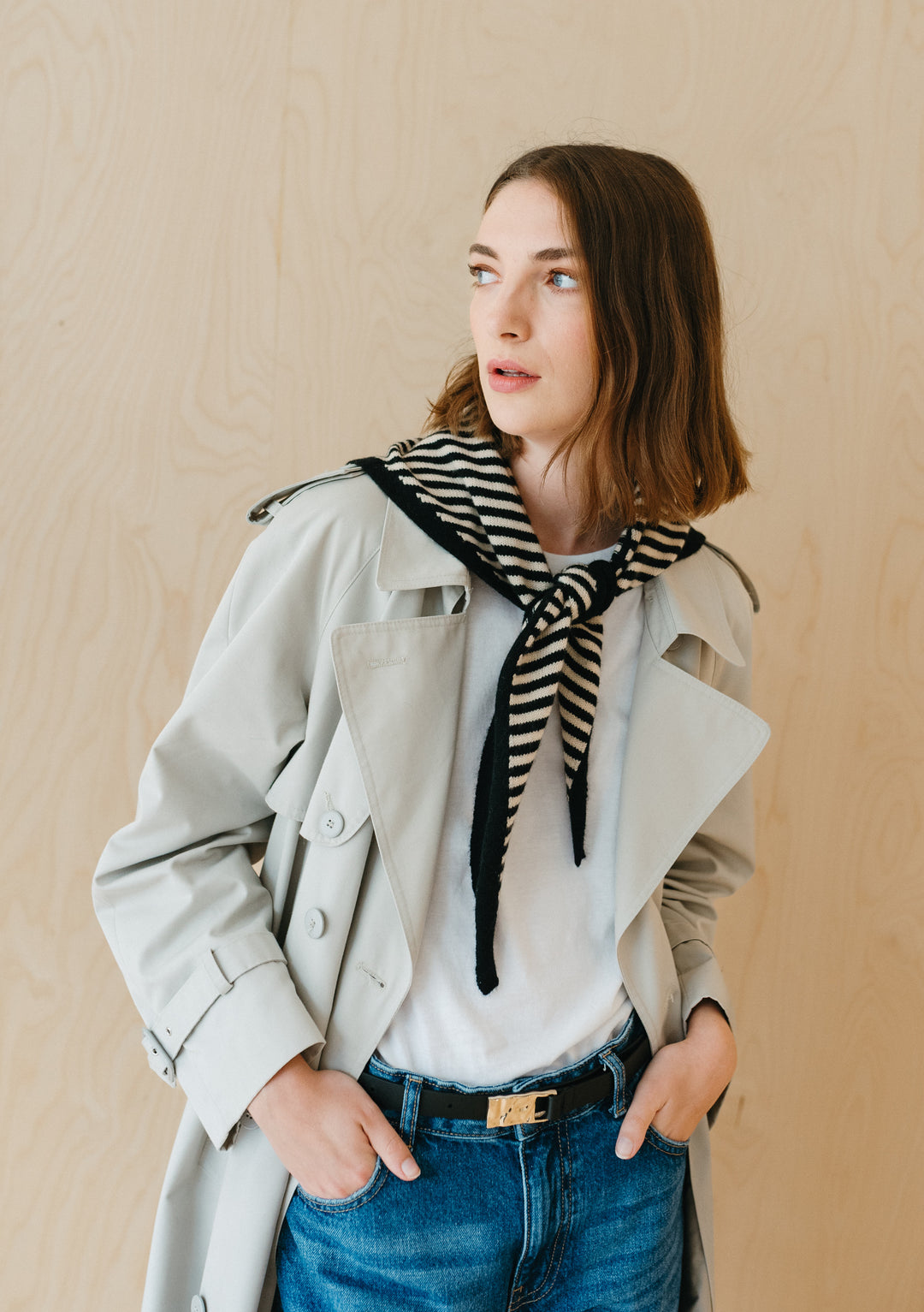 Person wearing TBCo Merino Triangle Scarf in Black Stripe styled casually with beige trench coat and blue denim against neutral wall