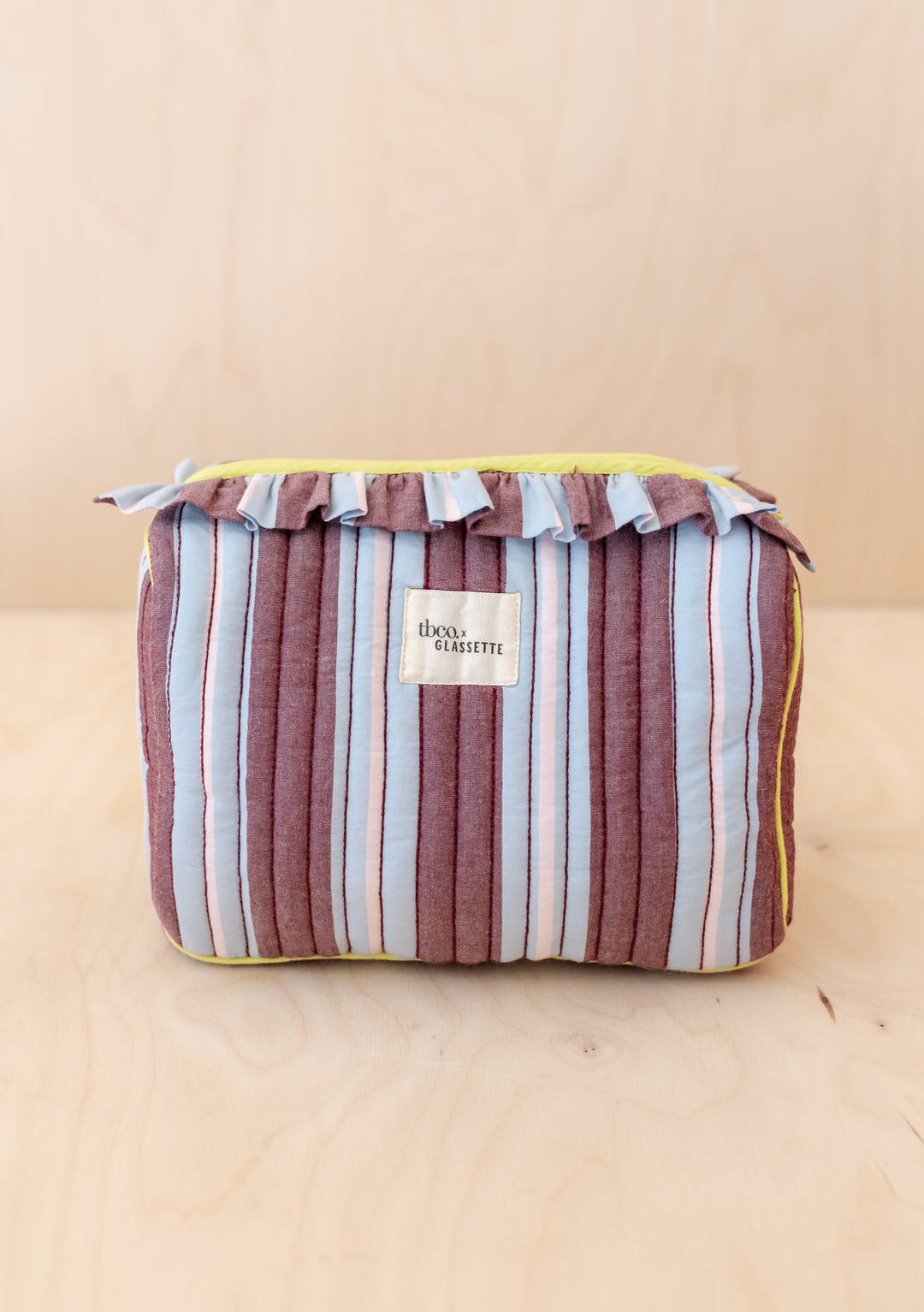 TBCo x Glassette Cotton Wash Bag in Berry Stripe, showcasing quilted vertical stripes, neon piping, ruffle trim, and a visible woven label on a wooden surface.