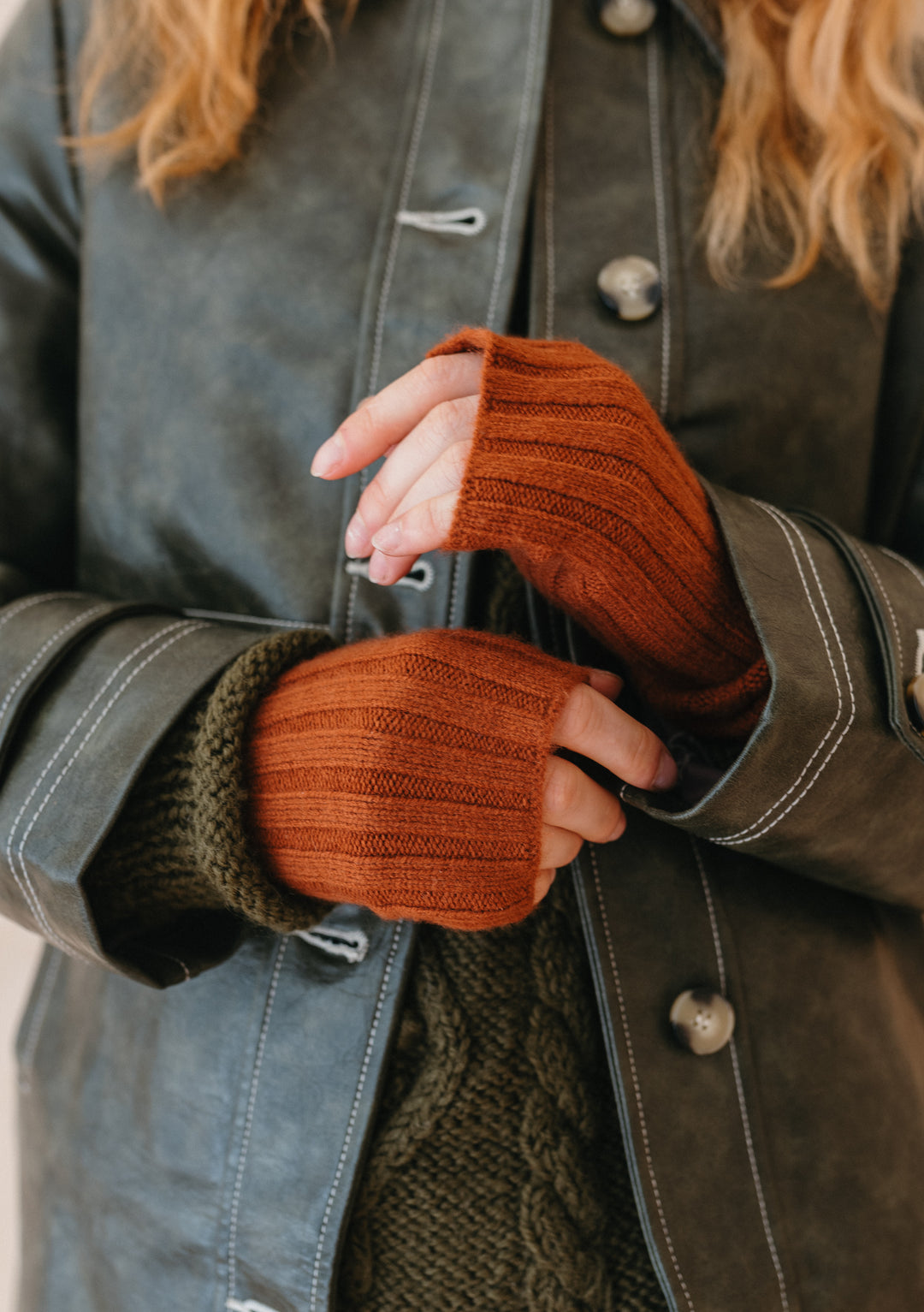 TBCo rust-coloured cashmere and merino wrist warmers worn with grey leather jacket showing ribbed knit texture and fingerless design