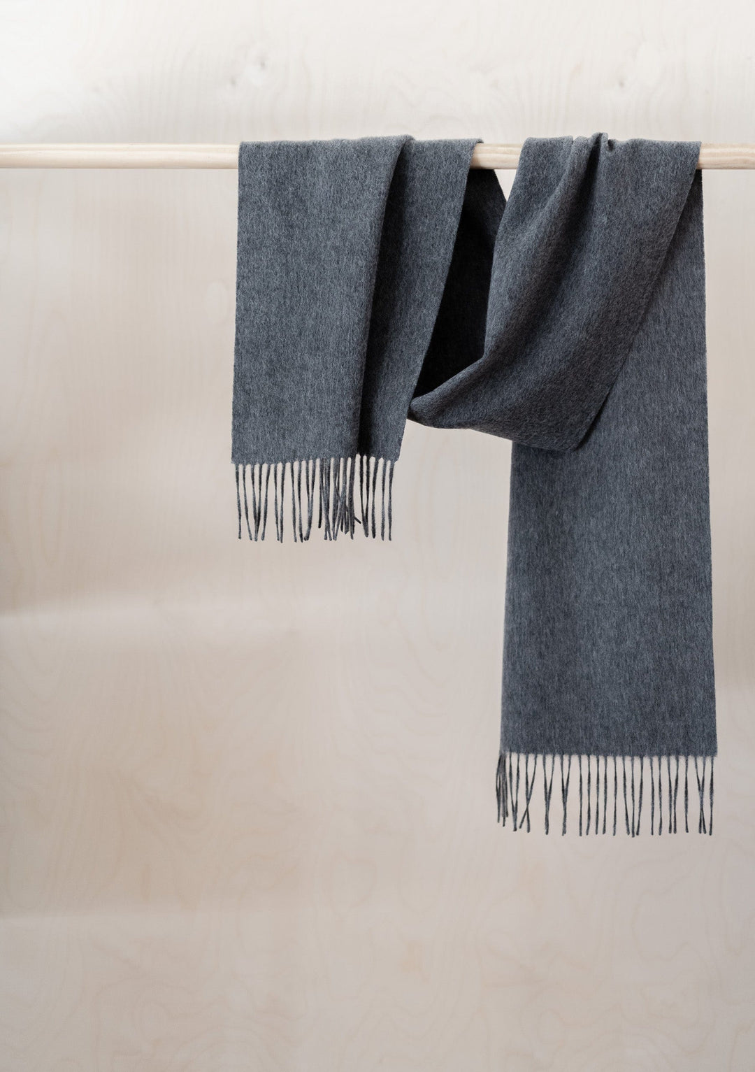 Luxurious TBCo charcoal melange cashmere scarf with black fringe detail, draped elegantly over wooden rail against neutral wall