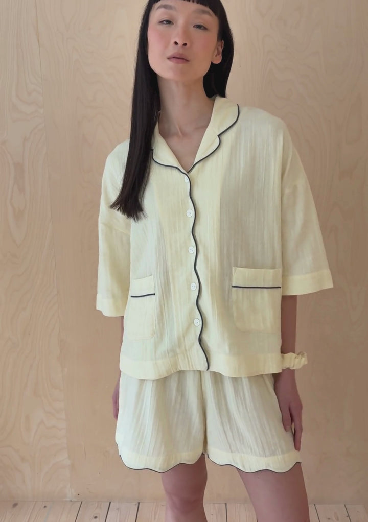 A model poses against a neutral ply backdrop, wearing a buttermilk yellow TBCo pyjama shorts and shirt set.
