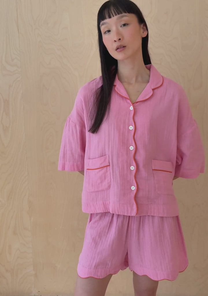 A model poses against a neutral ply backdrop, wearing a pink cotton TBCo pyjama shorts and shirt set.