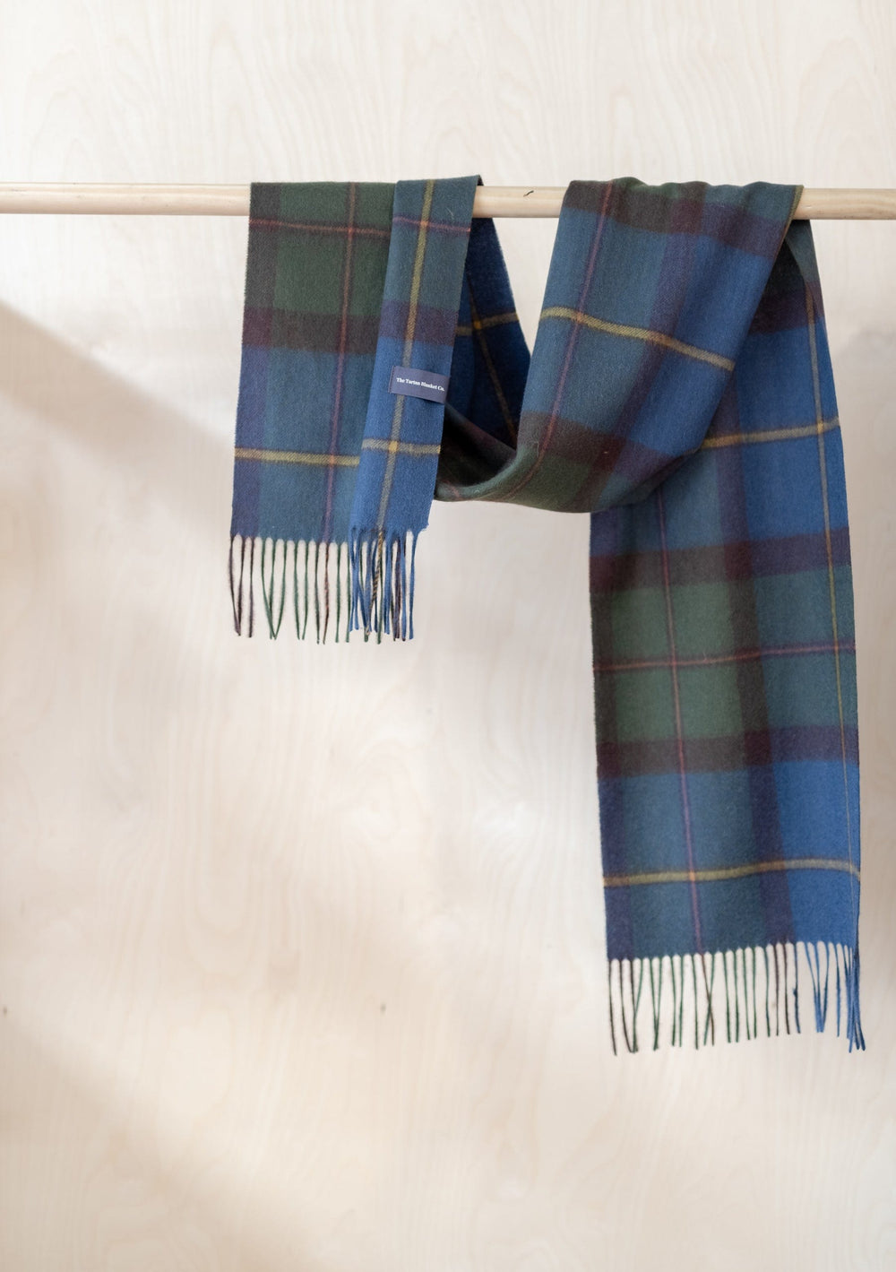 TBCo Macleod of Harris Antique Tartan lambswool scarf draped over wooden rail, showcasing blue and green plaid pattern with fringe