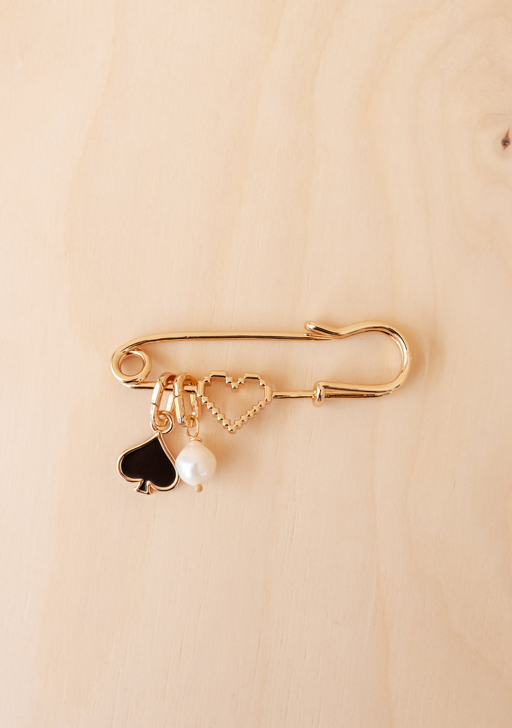 TBCo x frkl. Circus Scarf Pin: Oversized gold-tone safety pin with pixel heart, black spade, and pearl charms, celebrating bold, expressive styling.
