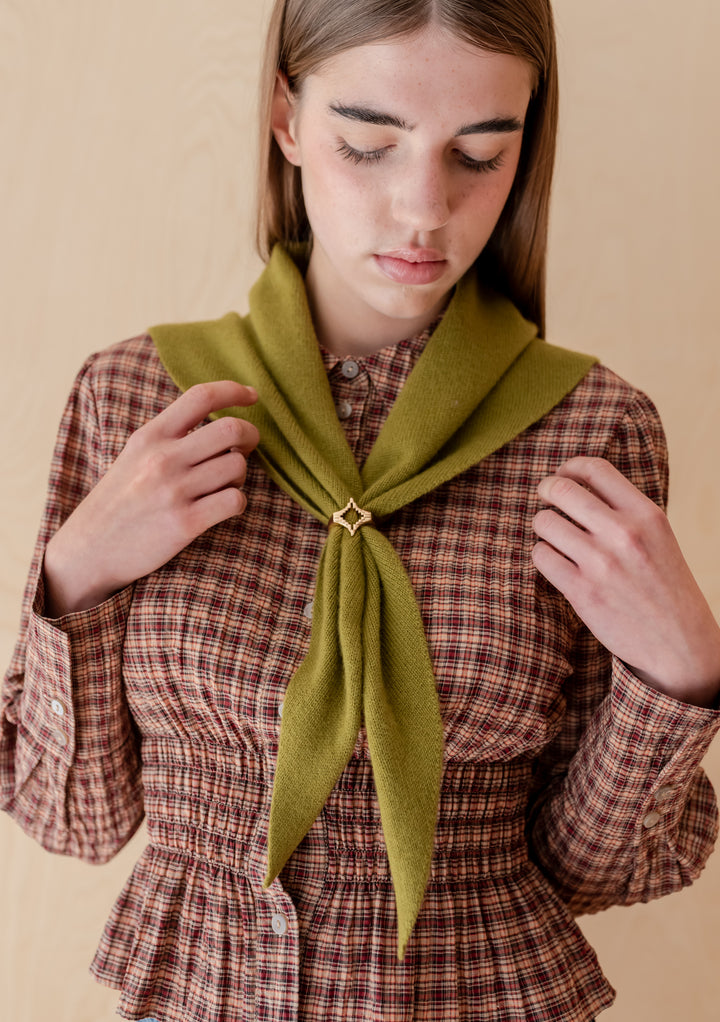 Young person wearing a red-brown blouse with an olive merino wool triangle scarf, featuring the TBCo x frkl. Diamond Scarf Ring, gold-tone star brooch securing the scarf.
