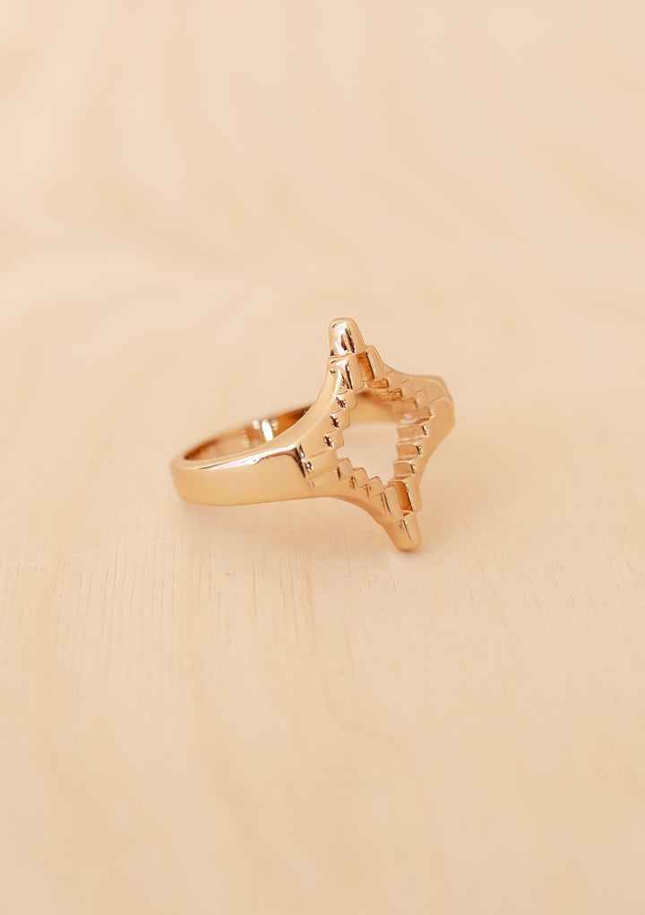 TBCo x frkl. Diamond Scarf Ring featuring a star-like, pixelated design in gold-toned brass, resting on a light wooden surface. Ideal for securing wool triangle scarves.